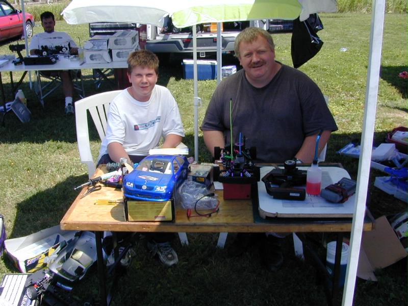 Man and teen at outdoor table with RC cars, fuel bottle, and tools at a hobby event