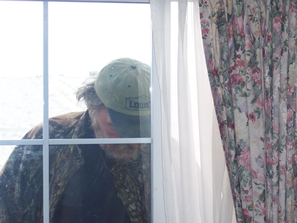 Man in Lowe's cap and camo jacket peers through window pane. Floral curtains visible