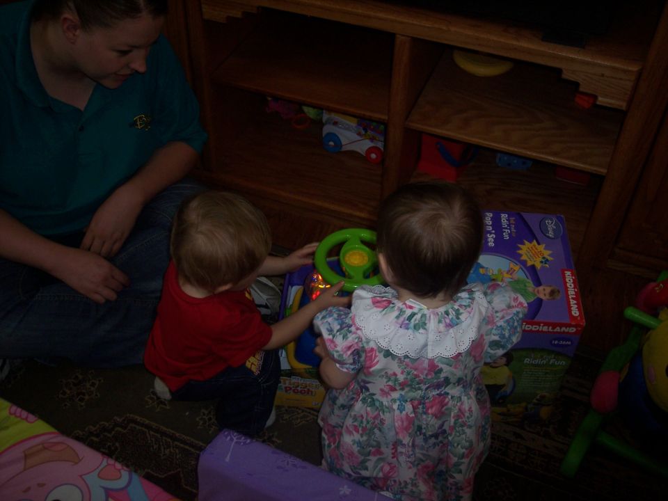Two toddlers play with a toy steering wheel and a KiddieLand Ridin' Fun box. A woman watches them