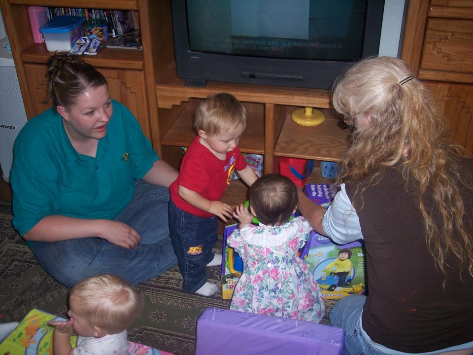 Four people, including two babies and two adults, are on the floor with toys in front of a television