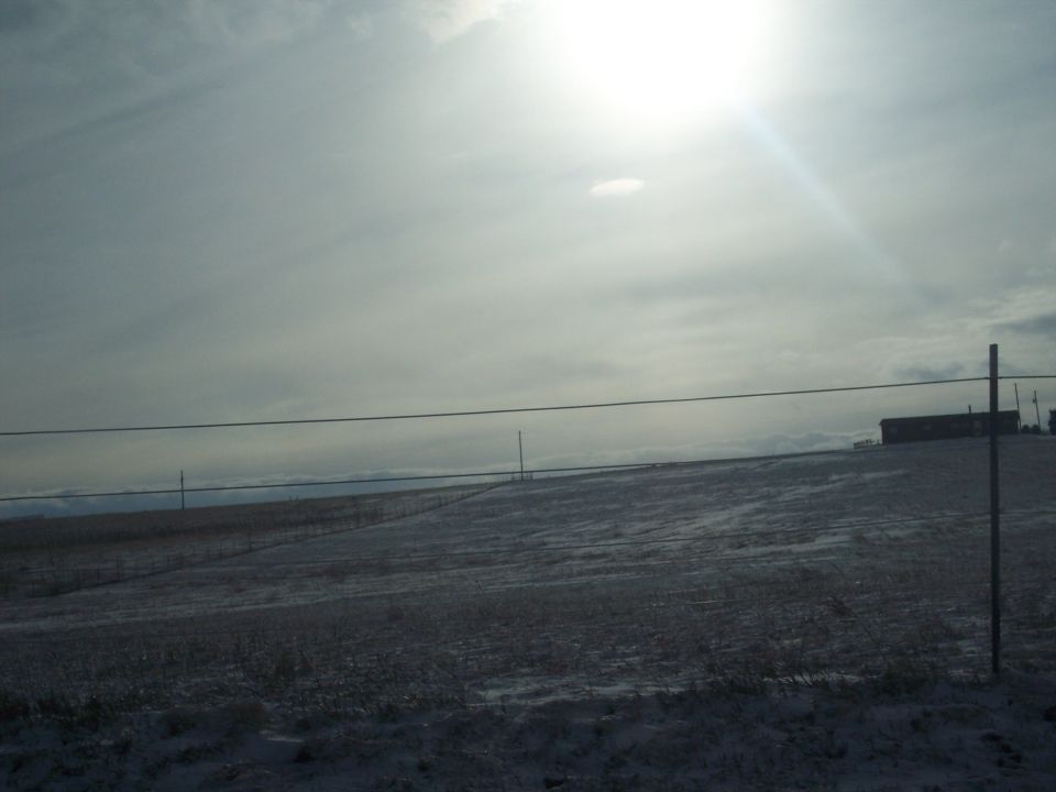 Snowy field under a bright, hazy sun with a distant building and power lines