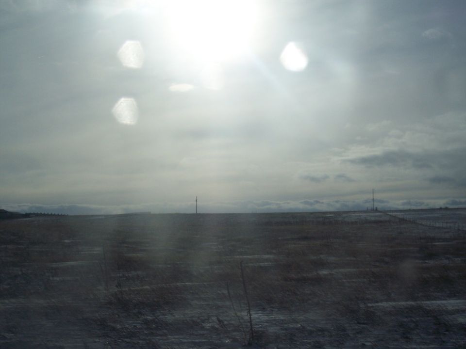 Wide shot of a desolate, snow-dusted field with a few distant radio towers under a bright, hazy sky