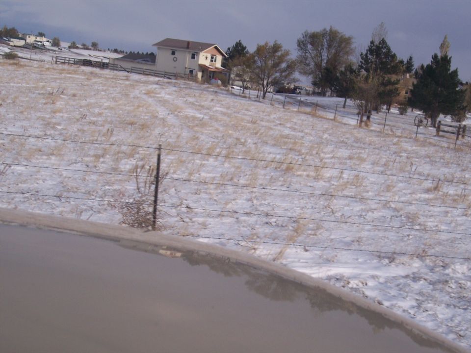 Snow covers a dry field with a house and trees in the background