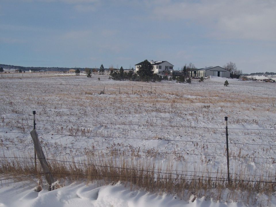 Snowy field with a house and barn in the distance under a cloudy sky
