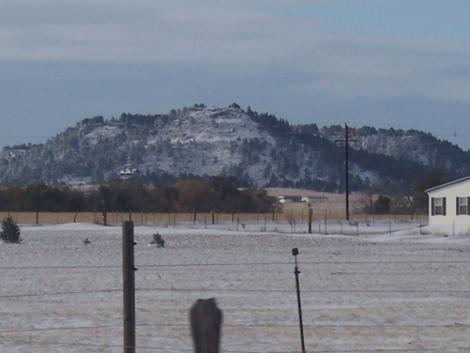 snow on hills and trees, with a house on the right and a fence in the foreground