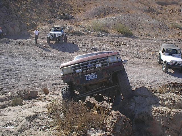 Faded red Ford pickup climbing a rock ledge nose-up on a desert trail with Jeeps waiting behind