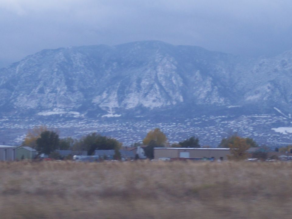 Snow-covered mountains loom over a distant town and dry grass field