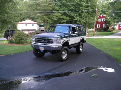 1994 Ford Bronco - Three-quarter front view of a lifted green-over-white full-size Ford Bronco parked on a freshly sealed asphalt driveway, wearing chrome-slo…