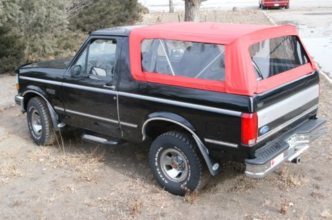 1994 Ford Bronco - Three-quarter rear view of a black full-size Ford Bronco fitted with a bright red aftermarket soft top in place of the factory fiberglass h…
