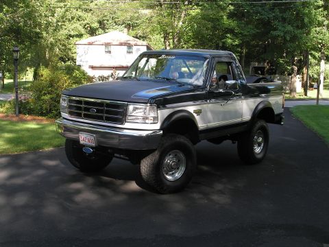 1994 Ford Bronco - A two-tone dark green and silver Ford Bronco full-size SUV, lifted with oversized tires and chrome wheels, parked on a paved driveway surro…