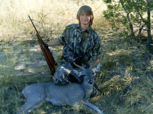 Hunter in camouflage with rifle poses over a downed deer in a dry, grassy field with sparse trees
