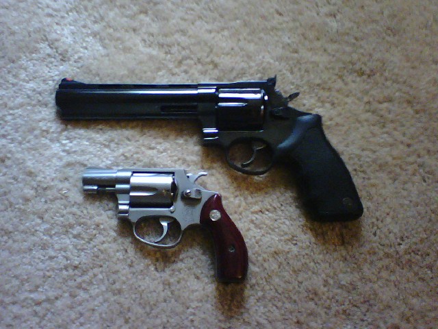 Two revolvers on a carpeted surface. One is a large black revolver with a long barrel, and the other is a smaller silver revolver with a dark red grip