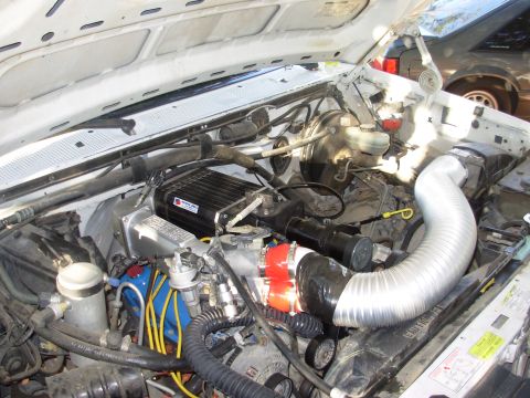 1994 Ford Bronco - The engine bay of a vehicle, an aftermarket air intake system and a black electronic box. Another vehicle is partially visible in the backg…