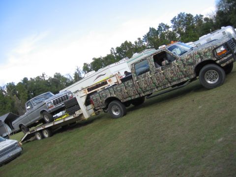 1979 Ford F-150 - A camouflaged Ford F-250 truck is seen from the side, towing a trailer carrying another pickup truck.