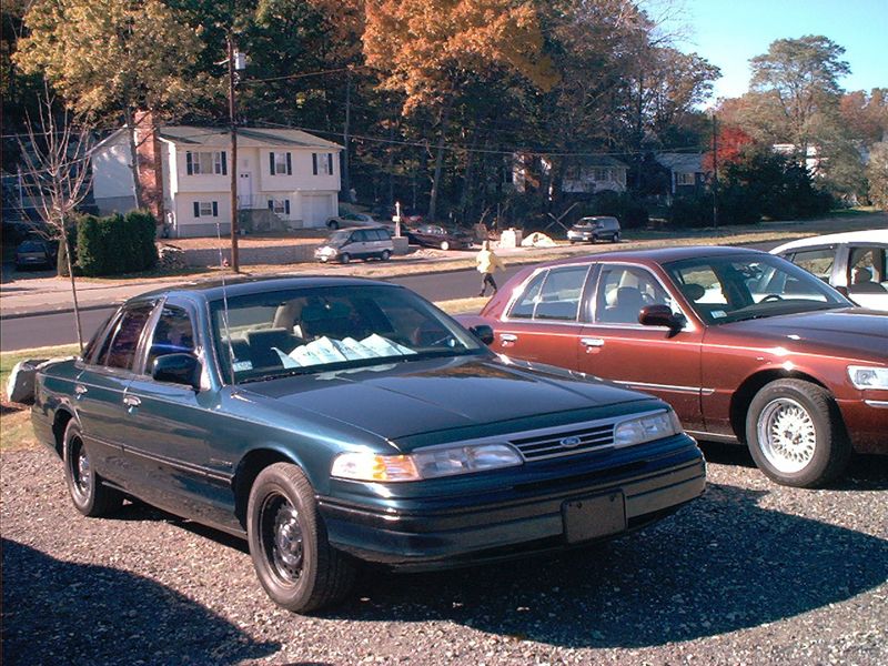 Green Ford Crown Victoria front three-quarter on gravel with maroon Mercury sedan beside