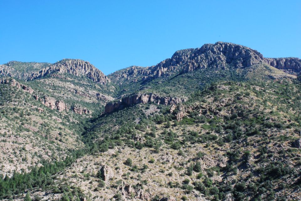 Rocky mountains with scattered trees under a clear blue sky. Watermark reads SUPER MOTORS