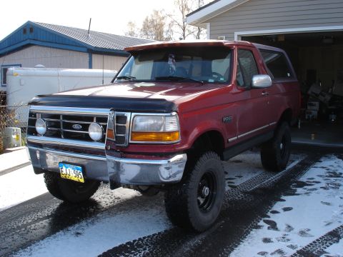 1996 Ford Bronco - This red Ford Bronco is parked outside a garage on a snowy day, showing its lifted stance and off-road tires.