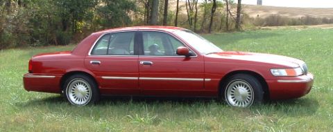 1999 Mercury Grand Marquis - A red 1999 Mercury Grand Marquis is parked in a grassy field with trees in the background.