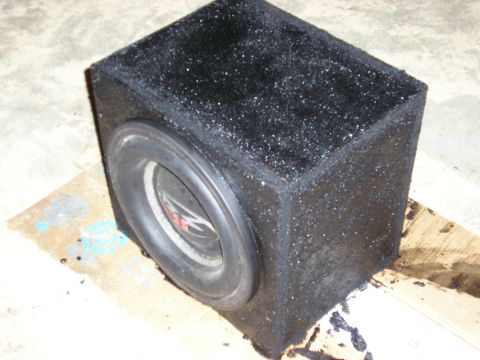 1996 Ford Bronco - A single black carpet-covered subwoofer enclosure sitting on cardboard on a concrete garage floor, with one round woofer mounted in the fro…