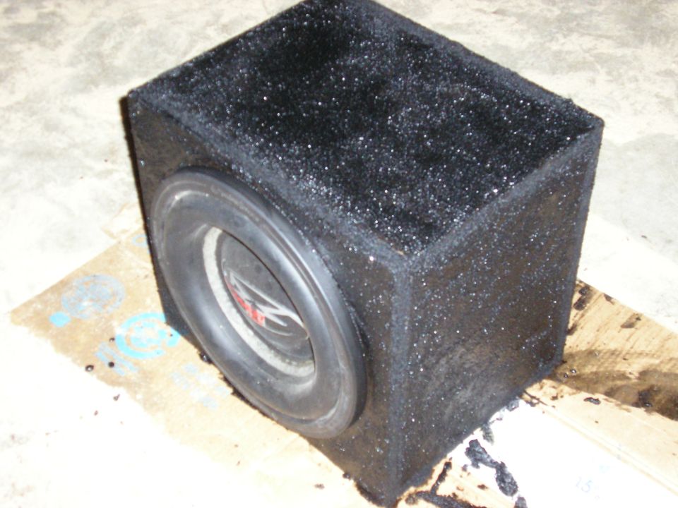 Black carpeted single-subwoofer box with rubber-surround woofer on a concrete garage floor