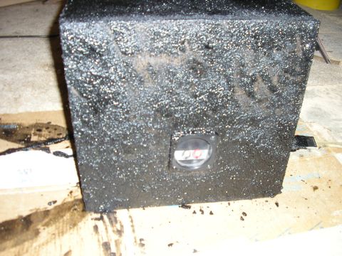 1996 Ford Bronco - A square subwoofer enclosure freshly coated in heavily textured black bedliner, still wet and sitting on cardboard masking on a garage floo…