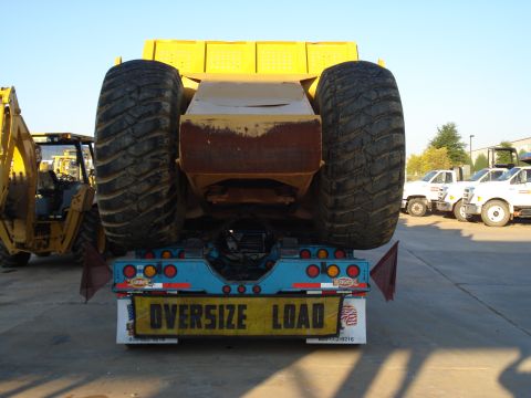 1966 International School Bus - A large piece of yellow heavy machinery is transported on a trailer, with other vehicles visible in the background.