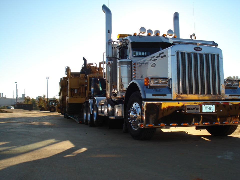 Silver Peterbilt semi-truck with lowboy trailer hauling yellow Caterpillar equipment