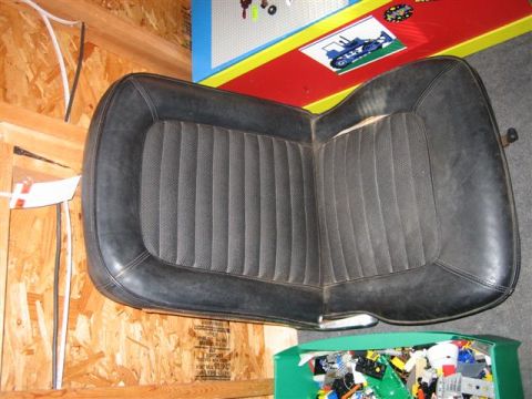 1900 Yugo Vega - A black car seat with a ribbed fabric insert is shown, with some text visible on a piece of wood in the background.