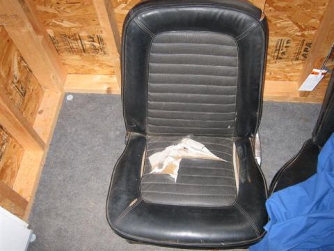 1900 Yugo Vega - A worn black car seat is shown in a room with unfinished wood walls and carpeted floor.