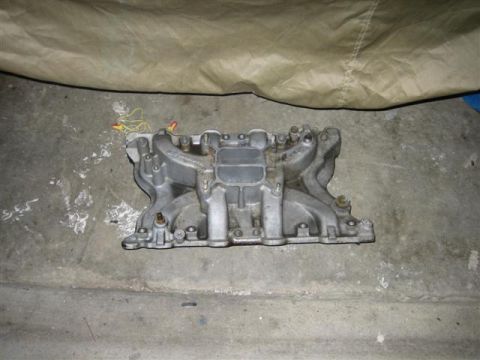 1900 Yugo Vega - An intake manifold rests on a concrete floor, partially covered by a tan tarp.