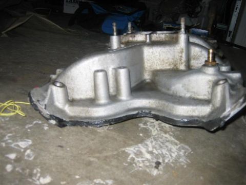 1900 Yugo Vega - A metal engine component sits on a concrete floor, showing signs of wear and gasket residue.