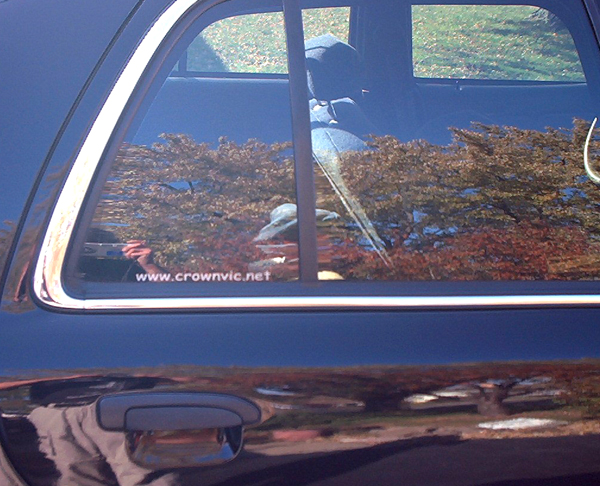 crownvic.net decal on rear door glass of dark blue sedan reflecting trees and a person