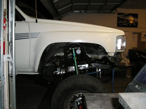 1986 Toyota Hilux - White pickup truck's front end is raised, revealing suspension modifications and a gauge in the wheel well.