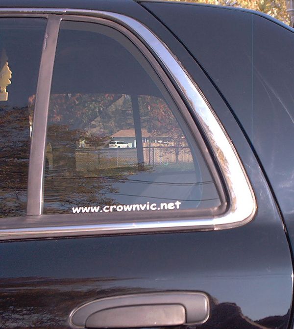 crownvic.net decal on rear quarter glass of dark sedan reflecting fenced yard and home
