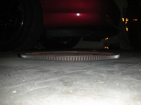 1992 Ford Crown Victoria - A close-up view of a ring gear resting on a concrete surface, with the underside of a red vehicle and a tire visible in the background.