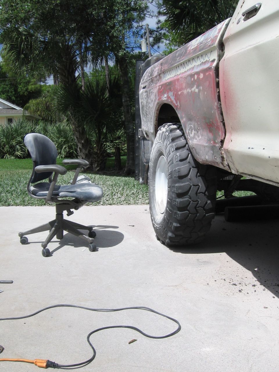 1979 Ford F-150 side view showing rear fender and large off-road tire. Body is in primer with some red showing through. Office chair sits nearby