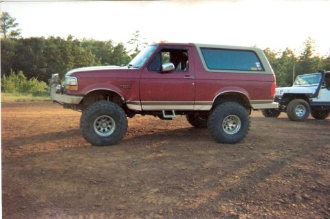 1995 Ford Bronco - A lifted red Ford Bronco is parked on a dirt road next to a white Jeep.
