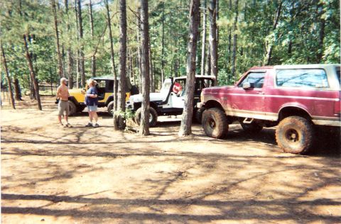 1995 Ford Bronco - A red Ford Bronco is parked in a wooded area alongside other off-road vehicles, with two people standing near a yellow Jeep.