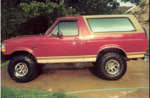 1995 Ford Bronco - Side view of a lifted red Ford Bronco with a tan top and gold trim, parked on a wet surface.