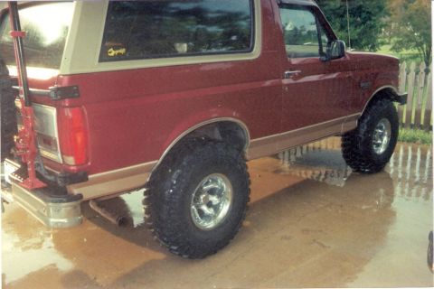 1995 Ford Bronco - A red Ford Bronco with tan trim and large off-road tires sits on a wet surface.