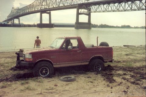 1995 Ford Bronco - A man stands by the water as a red 1995 Ford Bronco is stuck in the mud near a river.