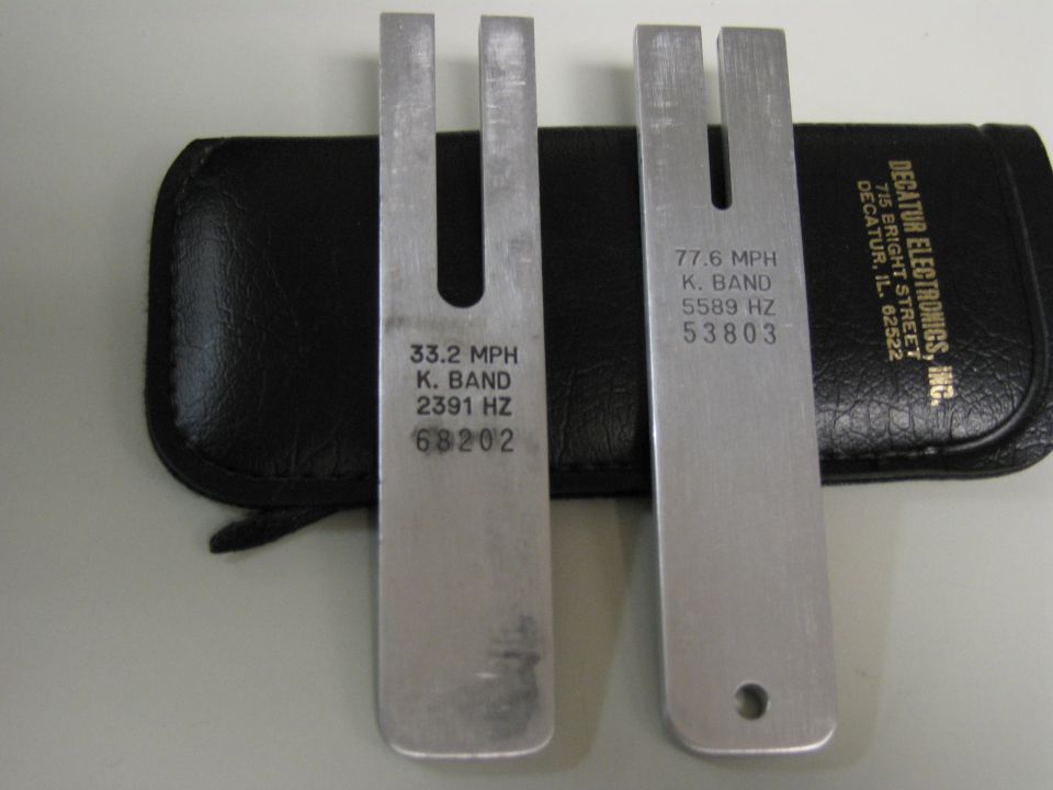 Two metal tuning forks with engraved text, one labeled 33.2 MPH K. BAND 2391 HZ 68202, the other 77.6 MPH K. BAND 5589 HZ 53803, next to a black case with Decatur Electronics Inc. text