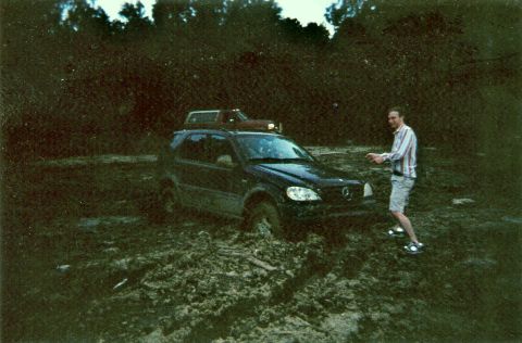 1995 Ford Bronco - A person stands near a dark SUV that is stuck in deep mud, with trees in the background.