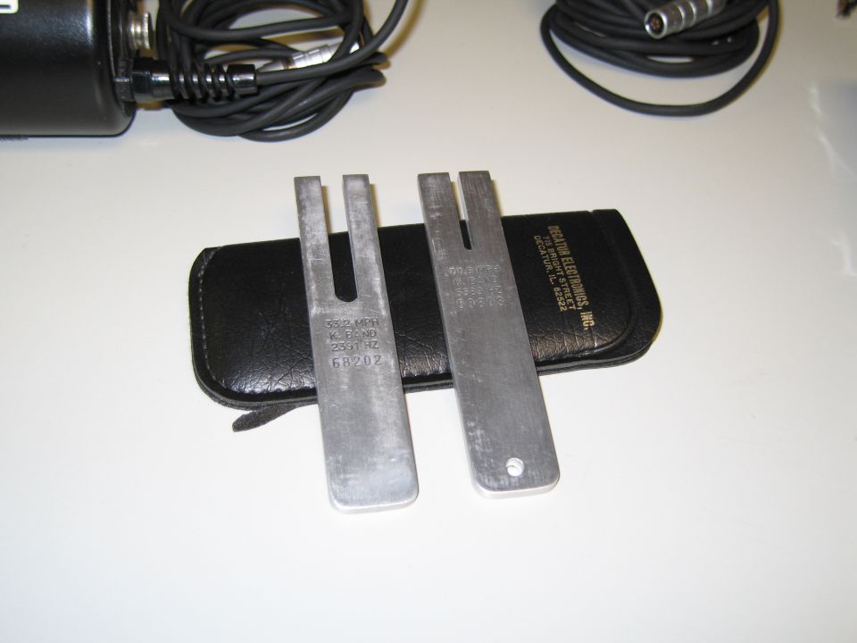 Two tuning forks with markings '332 MHZ K.B.ND 2331 HZ 68202' and '60.0 MHZ K.B.ND 2331 HZ 68202' resting on a black leather case next to an electronic device with cables