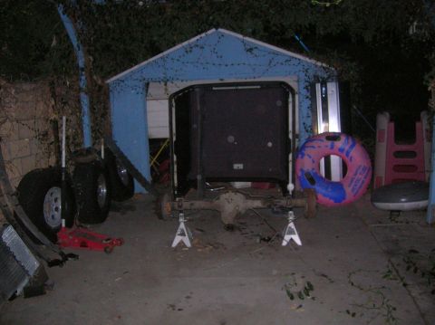 1985 Ford Bronco - Rear axle assembly supported by jack stands in a cluttered yard with a blue shed in the background.