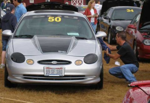 1997 Ford Taurus - Head-on view of a silver Ford Taurus SHO at a car show with a black cowl-style hood insert with pinstriping and an "SHO" decal, smoked head…