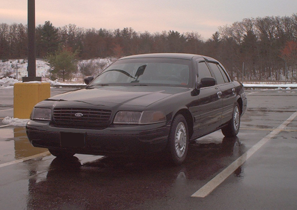 Black Ford Crown Victoria sedan parked in a wet lot with snow and bare trees behind