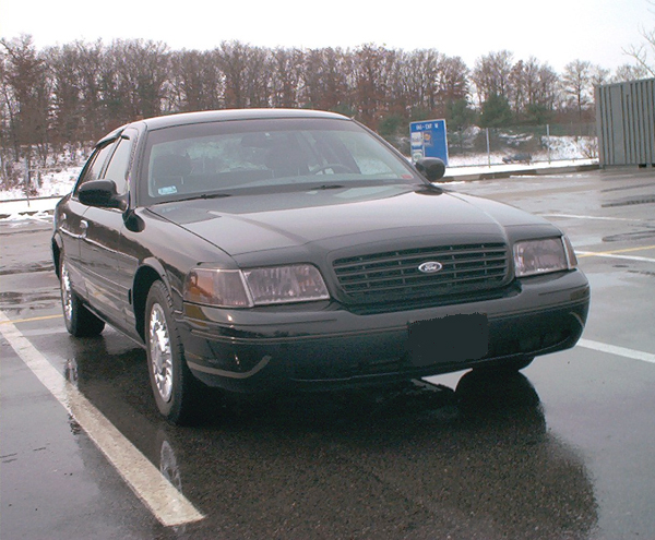 Black Ford Crown Victoria sedan in a wet parking lot with bare trees
