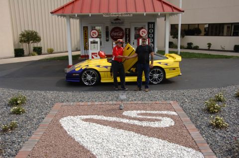 1989 Chevrolet Corvette - A yellow sports car with purple flames and a gullwing door open is displayed at a vintage gas station.