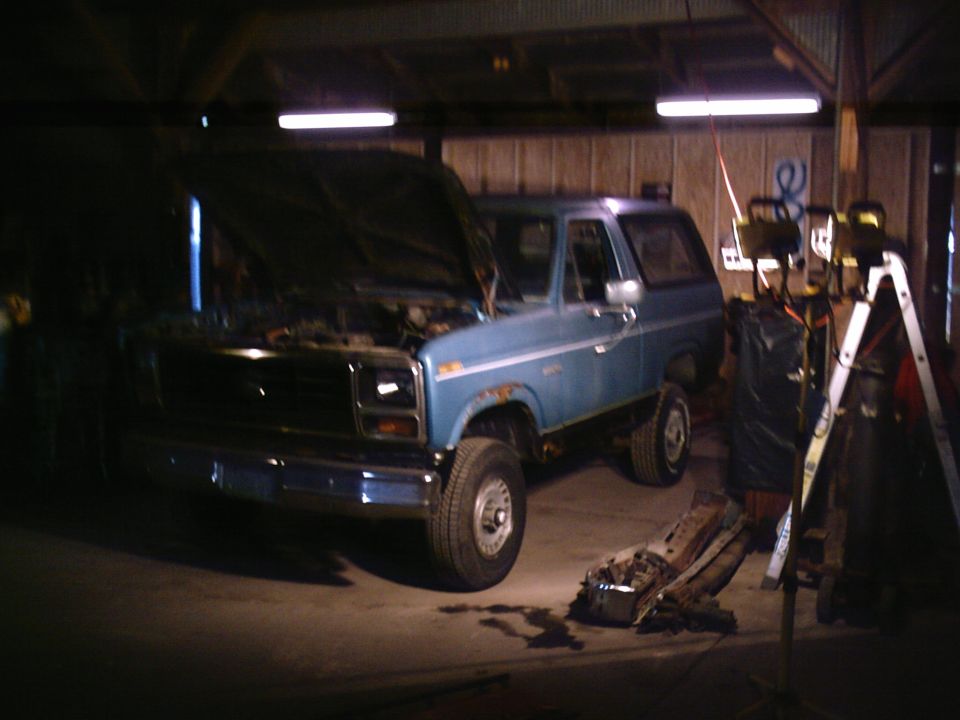 blue Ford Bronco with hood open in a garage, showing rust on the body